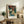 May include: A framed Japanese woodblock print featuring a dramatic coastal landscape with red autumn maple trees, white cliffs, and blue water displayed on a rustic wooden sideboard beside a fern plant, ceramic vase, books, and table lamp in a neutral-toned living room setting.