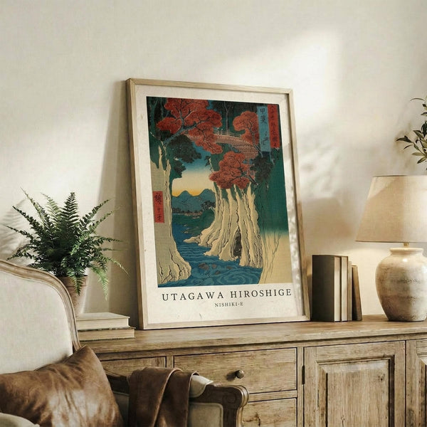 May include: A framed Japanese woodblock print featuring a dramatic coastal landscape with red autumn maple trees, white cliffs, and blue water displayed on a rustic wooden sideboard beside a fern plant, ceramic vase, books, and table lamp in a neutral-toned living room setting.