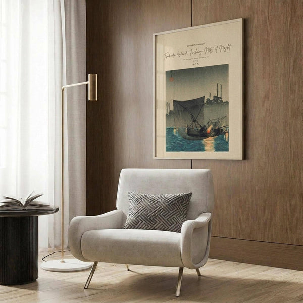May include: Japanese woodblock print poster featuring fishing boats at twilight with dark sails on blue water, displayed in light wooden frame on wood-paneled wall. Modern beige armchair with geometric pillow positioned below artwork next to brass floor lamp and dark side table with open book. Neutral interior with curtains and hardwood floors.