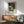 May include: Framed Japanese woodblock print showing autumn trees with red foliage beside a waterfall and blue water, displayed above a wooden sideboard with ceramic lamp. Mid-century modern green upholstered chair with wooden arms sits nearby. Warm sunlight creates shadows across white wall in minimalist room setting.