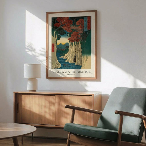 May include: Framed Japanese woodblock print showing autumn trees with red foliage beside a waterfall and blue water, displayed above a wooden sideboard with ceramic lamp. Mid-century modern green upholstered chair with wooden arms sits nearby. Warm sunlight creates shadows across white wall in minimalist room setting.
