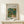 May include: Framed art print of Pierre-Auguste Renoir's impressionist painting showing two small figures walking down a tree-lined path. The artwork features soft brushstrokes in greens, blues, and warm earth tones depicting a lush garden scene. The print has white matting in a thin wooden frame, displayed on a light wood surface alongside a small white gift box with natural twine and evergreen sprig against a cream wall.