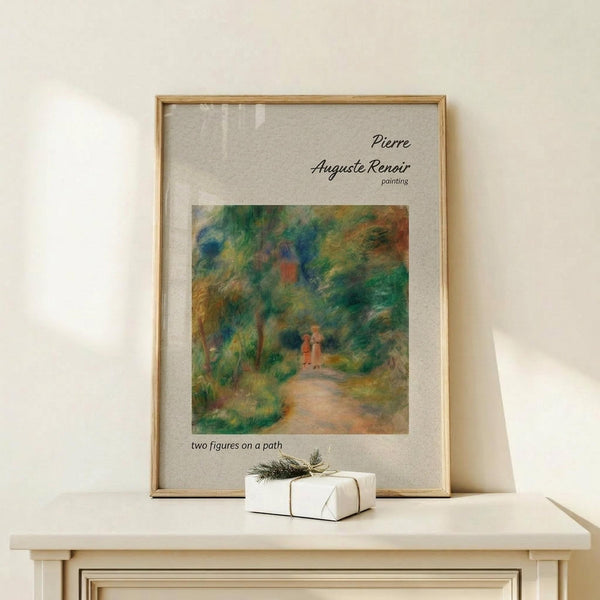 May include: Framed art print of Pierre-Auguste Renoir's impressionist painting showing two small figures walking down a tree-lined path. The artwork features soft brushstrokes in greens, blues, and warm earth tones depicting a lush garden scene. The print has white matting in a thin wooden frame, displayed on a light wood surface alongside a small white gift box with natural twine and evergreen sprig against a cream wall.