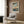May include: Japanese woodblock print poster in light wood frame showing autumn maple branches with orange leaves over a blue river landscape. Mounted on wood-paneled wall above beige upholstered armchair with geometric patterned pillow. Modern reading nook with brass floor lamp and dark round side table holding open book near window with white curtains.
