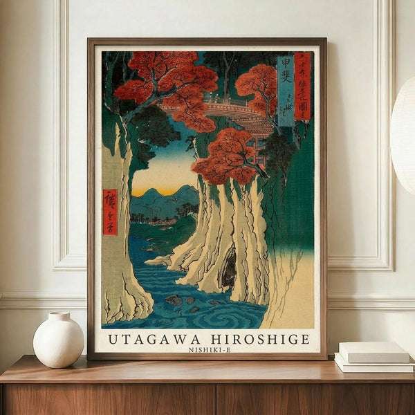 May include: Framed Japanese woodblock print featuring autumn maple trees with vibrant red-orange foliage overlooking a turquoise waterfall cascading down white limestone cliffs. The traditional ukiyo-e artwork shows distant mountains and includes Japanese text cartouches. The brown-framed poster sits on a wooden sideboard next to a white ceramic vase and books in a bright room with white walls and decorative molding.