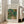 May include: Framed Renoir impressionist painting print showing two figures walking on a garden path surrounded by lush green foliage with touches of blue, orange and yellow. The artwork is displayed in a light wood frame with white matting, leaning against a beige armchair in a bright living room with large windows and wooden floors.