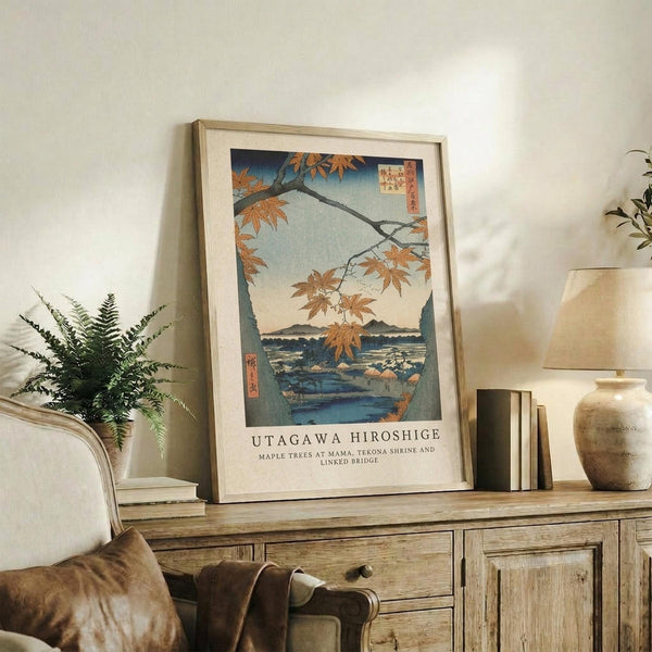 May include: Framed Japanese woodblock print featuring autumn maple trees with orange leaves overlooking a blue water landscape, displayed on rustic wooden sideboard alongside fern plant, ceramic vase, and books in neutral-toned living room setting