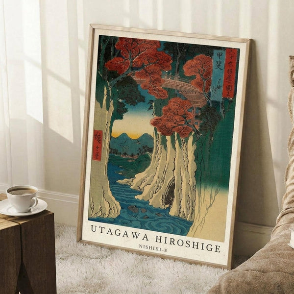 May include: Framed Japanese woodblock print showing autumn trees with red foliage above white cliffs and blue water, with mountains in the distance. The artwork features traditional ukiyo-e style with Japanese text cartouches. Print displays in light wood frame leaning against white wall beside wooden side table with coffee cup and cream fabric.