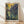 May include: A framed Van Gogh-style poster featuring purple irises with flowing green stems against a golden yellow background, displayed in a light wood frame on wooden flooring against a white wall with natural sunlight casting diagonal shadows across the scene.