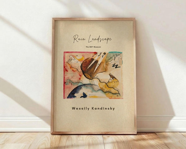 May include: Framed art print featuring Wassily Kandinsky's abstract Rain Landscape painting with flowing organic forms in red, yellow, blue, and earth tones against cream background. Wooden frame leans against white wall on light wood floor with soft natural lighting creating diagonal shadows.