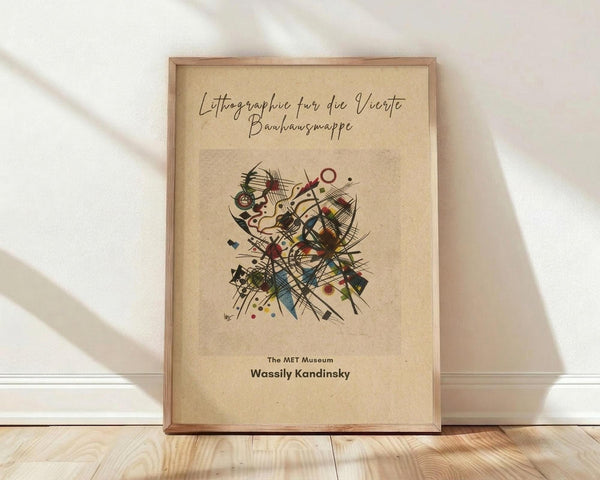 May include: Framed vintage-style poster with beige background featuring abstract Kandinsky artwork with colorful geometric shapes, lines, and forms in red, blue, black, and earth tones. German text at top, "The MET Museum" and "Wassily Kandinsky" at bottom. Natural wood frame leaning against white wall on light wooden floor with soft window lighting casting shadows.