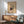 May include: Abstract modernist art poster in wood frame hanging above mid-century sideboard with white ceramic lamp. Mint green upholstered armchair with wooden arms sits beside round coffee table. Warm sunlight casts geometric shadows across white wall. Artwork features colorful geometric shapes, circles, and lines in red, blue, yellow, and black on beige background.
