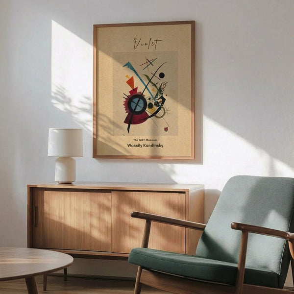 May include: Abstract modernist art poster in wood frame hanging above mid-century sideboard with white ceramic lamp. Mint green upholstered armchair with wooden arms sits beside round coffee table. Warm sunlight casts geometric shadows across white wall. Artwork features colorful geometric shapes, circles, and lines in red, blue, yellow, and black on beige background.