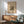 May include: Framed abstract art poster with colorful geometric shapes, lines and forms in Kandinsky's distinctive style, displayed above a wooden sideboard with white ceramic lamp. Modern green upholstered armchair with wooden arms sits nearby on wooden flooring. Warm natural sunlight creates shadows across white wall, showcasing mid-century modern interior design aesthetic.