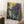 May include: Van Gogh's Irises reproduction poster in wooden frame, leaning against white wall in cozy room setting. Artwork features vibrant purple-blue iris flowers with flowing green stems against golden yellow swirled background. Poster has white border with artist name at bottom. Scene includes white coffee cup on wooden block, beige knit throw, and white textured rug on wooden floor with natural window light.
