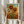 May include: Hands holding a framed art print of vibrant sunflowers in Van Gogh's distinctive style, featuring golden yellow blooms with dark centers against a blue sky and textured field background. The print has a light wood frame with "Vincent van Gogh" text below the image. Dried wheat stalks visible to the left, neutral beige clothing and soft curtains in background create a warm, natural styling scene.