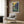 May include: Van Gogh's Irises poster in light wooden frame hanging on warm wood-paneled wall above beige upholstered armchair. Artwork shows purple irises with green stems against yellow background. Scene includes brass floor lamp, black side table with open book, geometric patterned throw pillow, and light curtains by window.