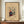 May include: Abstract modernist artwork in wooden frame on wooden sideboard next to white ceramic vase and books. The framed piece features geometric shapes, lines, and forms in vibrant colors including teal, red, yellow, and purple on beige background. Museum-style poster with text attribution below the colorful abstract composition. Interior setting shows neutral walls with white trim molding.