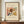 May include: Framed poster featuring Kandinsky's abstract watercolor painting with flowing organic forms in browns, yellows, blues and reds on cream background. Modern wood frame displayed on wooden sideboard next to white ceramic vase and stacked books against white wainscoted wall.