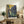 May include: Van Gogh-style poster print of blue and purple irises with flowing stems against a golden yellow sky background, displayed in thin wooden frame on rustic wood sideboard alongside fern plant, ceramic vase, stacked books, and cream upholstered chair in warmly lit living room setting.