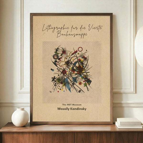 May include: Framed vintage-style art print featuring Wassily Kandinsky's abstract composition with colorful geometric shapes, lines, and forms on cream background. German text at top reads "Lithographie fur die Vierte Bauhausmappe" with "The MET Museum" and artist name below. Brown wooden frame displayed on walnut credenza beside white ceramic vase and stacked books against cream wainscoted wall.