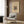 May include: Framed beige poster with abstract geometric artwork featuring colorful intersecting lines and shapes, displayed on wood-paneled wall above light gray upholstered armchair with geometric patterned throw pillow, alongside brass floor lamp and black side table with open book in modern neutral-toned room setting.