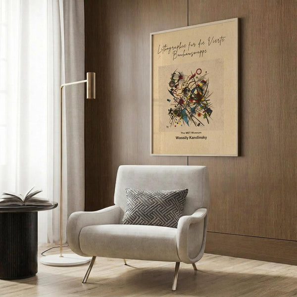 May include: Framed beige poster with abstract geometric artwork featuring colorful intersecting lines and shapes, displayed on wood-paneled wall above light gray upholstered armchair with geometric patterned throw pillow, alongside brass floor lamp and black side table with open book in modern neutral-toned room setting.