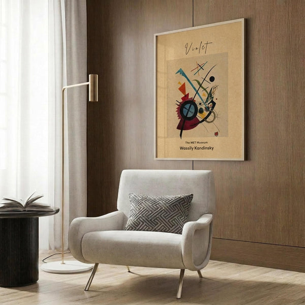 May include: Framed Kandinsky abstract art print with colorful geometric shapes on beige background, displayed on wood paneled wall above beige upholstered armchair with patterned pillow. Modern brass floor lamp and dark round side table with open book visible in neutral-toned living room setting.