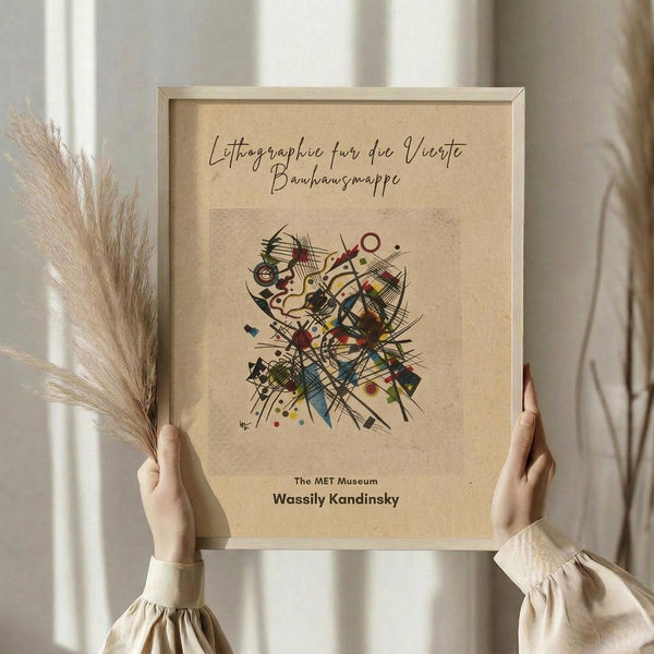 May include: Hands holding a framed abstract art print featuring Kandinsky's colorful geometric composition with intersecting lines, circles, and angular shapes in red, blue, yellow, and black on beige background. Light wood frame, pampas grass visible to left, neutral cream curtains behind, soft natural lighting.