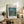 May include: Framed Van Gogh museum poster featuring Starry Night Over the Rhone painting with swirling blue night sky, golden stars, and water reflections. Light wood frame displayed on rustic wooden sideboard alongside fern plant, ceramic vase, books, and beige upholstered chair in cozy neutral-toned living room setting.