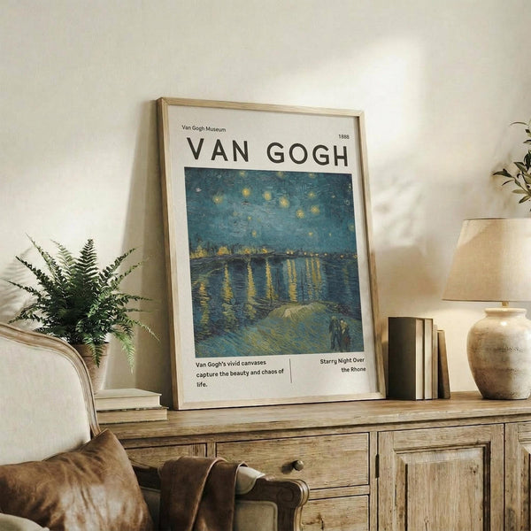 May include: Framed Van Gogh museum poster featuring Starry Night Over the Rhone painting with swirling blue night sky, golden stars, and water reflections. Light wood frame displayed on rustic wooden sideboard alongside fern plant, ceramic vase, books, and beige upholstered chair in cozy neutral-toned living room setting.