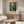 May include: A framed poster showing a figure in a boat on water beneath a swirling sun and sky in Van Gogh's distinctive style, displayed above a walnut wood credenza in a modern living room with cream walls, white upholstered chair, glass coffee table, and brass floor lamp.