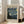 May include: Framed museum-style poster featuring Van Gogh's Starry Night painting with swirling blue sky, yellow stars, and dark cypress tree. The beige poster has "VAN GOGH" text at top and "Van Gogh Museum 1889" details. Light wooden frame leans against white wall next to beige upholstered chair in bright, minimalist living room with large windows and natural light casting shadows on hardwood floors.