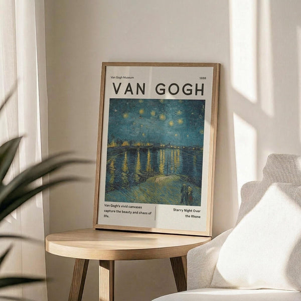May include: Framed Van Gogh museum poster featuring Starry Night Over the Rhone painting with swirling blue night sky and yellow stars reflecting on water. Displayed on round wooden side table next to beige upholstered chair with white pillow. Soft natural lighting creates shadows on white wall, with green plant leaves visible in corner.