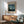 May include: Van Gogh museum poster featuring The Starry Night painting with swirling blue sky and yellow stars, displayed in thin wooden frame above natural wood sideboard. Mid-century modern living room with sage green upholstered chair, white ceramic table lamp, and warm sunlight casting geometric shadows on white wall.