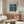 May include: Van Gogh museum poster featuring Starry Night Over the Rhone painting with swirling blue night sky, yellow stars, and water reflections, displayed in thin brown frame above wooden sideboard with sliding doors. Modern living room setting with white curved chair, round glass coffee table, brass floor lamp, and white table lamp on credenza.