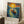 May include: Framed poster print featuring Van Gogh-inspired artwork showing a figure in a wooden boat on swirling blue water beneath a dramatic yellow sun and starry sky, displayed leaning against white wall corner next to coffee cup on wooden block and beige throw blanket on white furry rug