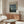 May include: Van Gogh Starry Night museum poster in thin wooden frame hanging above walnut credenza in minimalist living room. Poster shows swirling blue night sky with yellow stars and moon over village. Room features cream upholstered chair, glass oval coffee table, copper floor lamp, and white table lamp on credenza. Natural lighting creates warm shadows on beige walls.