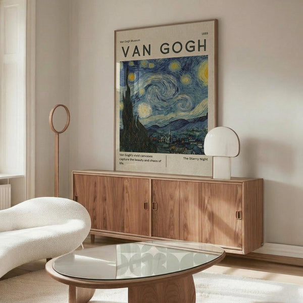 May include: Van Gogh Starry Night museum poster in thin wooden frame hanging above walnut credenza in minimalist living room. Poster shows swirling blue night sky with yellow stars and moon over village. Room features cream upholstered chair, glass oval coffee table, copper floor lamp, and white table lamp on credenza. Natural lighting creates warm shadows on beige walls.
