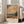 May include: Framed Van Gogh-inspired landscape poster featuring swirling orange and yellow sky over rolling hills with dark cypress trees and distant mountains. The print shows thick, textured brushstrokes in warm earth tones with French text below the artwork. White frame leans against beige sofa in bright room with tall windows and hardwood floors.