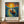 May include: Framed poster displayed on wooden console table featuring Van Gogh-inspired artwork of figure in small boat on swirling blue water with bright yellow sun and colorful sky. Text below reads "Vincent Van Gogh" with French description. White ceramic vase and books flank the brown-framed print against cream wainscoted wall.