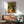 May include: Vincent van Gogh's olive trees painting print in thin wooden frame hanging on white wall above natural wood sideboard with white table lamp. Mint green mid-century armchair with wooden arms sits nearby. Artwork shows gnarled olive tree with golden-yellow foliage along winding path leading to small house, painted in van Gogh's characteristic swirling brushstroke style with vibrant yellows, greens, and earth tones. Warm sunlight streams across minimalist room.