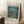 May include: Van Gogh museum-style poster in light wood frame leaning against white wall corner. Shows Starry Night Over the Rhone painting with swirling blue night sky, yellow stars, bridge over water with golden reflections, and two figures on shoreline. White border with black text and 1888 date. Coffee cup on wooden block and beige blanket nearby on textured white rug.