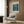 May include: Framed Van Gogh museum poster featuring Starry Night Over the Rhone painting with swirling blue night sky, golden stars reflecting on water, and figures on shoreline. Displayed above modern beige armchair with geometric pillow in contemporary living room with wood paneling, gold floor lamp, and side table with open book.