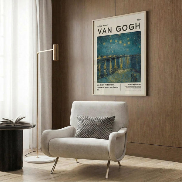 May include: Framed Van Gogh museum poster featuring Starry Night Over the Rhone painting with swirling blue night sky, golden stars reflecting on water, and figures on shoreline. Displayed above modern beige armchair with geometric pillow in contemporary living room with wood paneling, gold floor lamp, and side table with open book.
