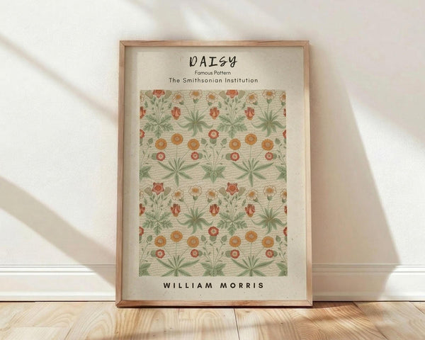 May include: Framed botanical print featuring William Morris Daisy pattern with repeating orange and coral flowers, green foliage on cream background. Natural wood frame displayed against white wall on light wooden floor with soft natural lighting creating diagonal shadows.