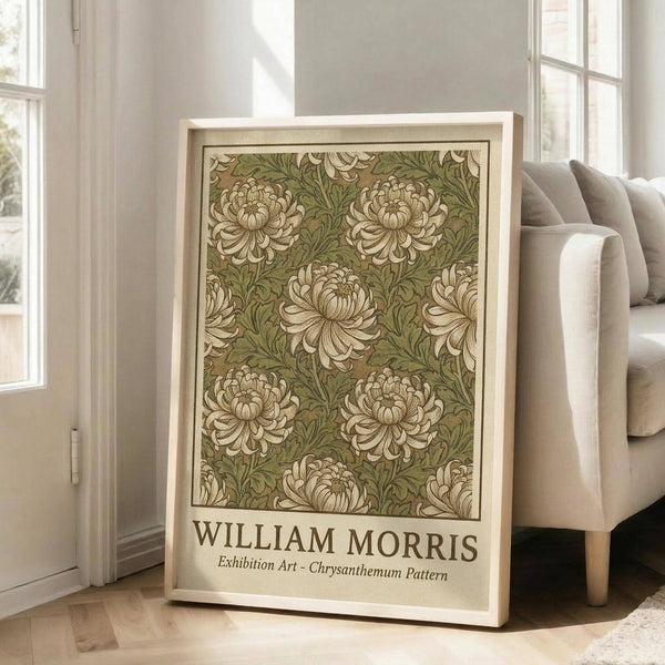 May include: Framed vintage-style botanical poster featuring William Morris chrysanthemum pattern in sage green and cream colors, displayed in bright living room with beige sofa, white walls, and natural light from tall windows. The artwork shows repeating floral motifs in Arts and Crafts style within light wood frame, leaning against white baseboard on hardwood floors.