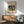 May include: Framed art print featuring Kandinsky's abstract waterfall painting with vibrant blues, greens, reds, and yellows displayed above a wooden sideboard. Mid-century modern living room setting with mint green upholstered chair, white table lamp, round wooden coffee table, and natural sunlight casting geometric shadows on white wall.