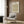 May include: Framed botanical poster with green foliage and orange circular flower motifs displayed on wood-paneled wall above beige upholstered armchair with geometric patterned pillow. Modern brass floor lamp with white circular base stands nearby, with dark round side table holding open book. Neutral tones throughout minimalist interior setting with natural lighting from window.