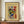 May include: Framed abstract art poster displayed on wooden sideboard next to white ceramic vase and stacked books. The artwork features vibrant swirling brushstrokes in green, blue, red, yellow and orange on cream background. Brown wooden frame complements warm neutral wall setting. Exhibition-style poster format with museum text above and artist name below the colorful abstract composition.
